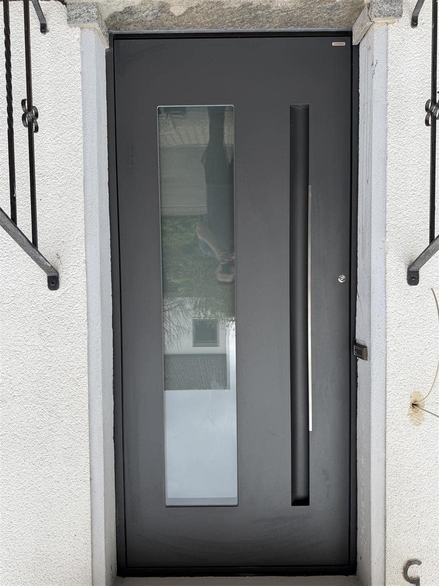 Moderne schwarze Haustür mit Glasausschnitt und vertikalem Stoßgriff, fertig montiert zwischen Geländer und Mauerwerk.